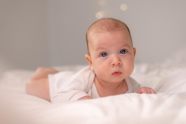 Bebê com olhos azuis em uma roupa branca está de barriga no estilo de vida de roupa de cama branca