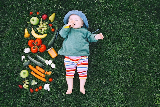 Bebê com roupas coloridas, experimentando comida e moldura de diferentes frutas frescas vegetais na grama verde