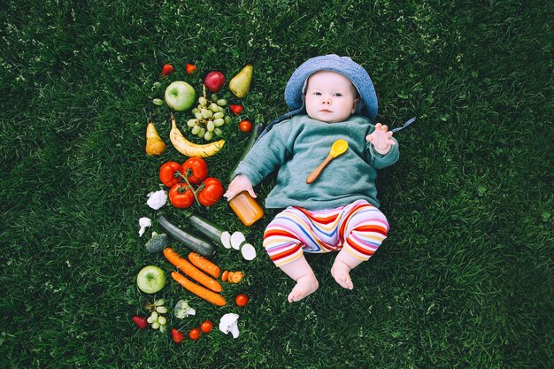 Bebê com roupas coloridas, experimentando comida e moldura de diferentes frutas frescas vegetais na grama verde