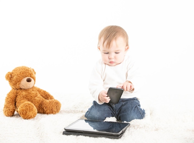 Bebê com um celular na mão e um tablet e um ursinho de pelúcia no chão