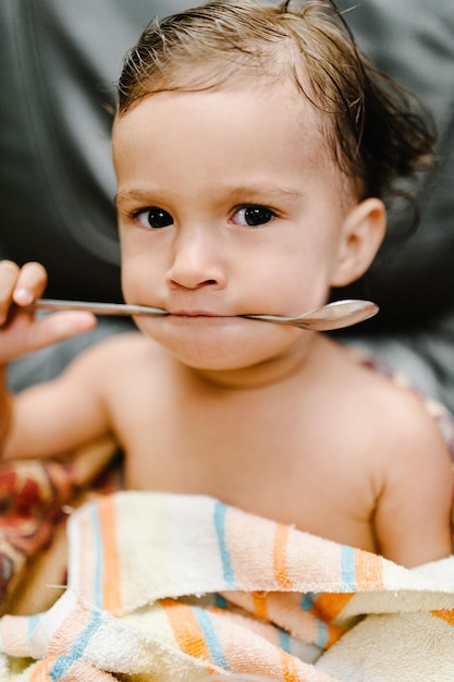 El bebé come una cuchara y se cubre con una toalla sentado en el sofá de la cama de los padres después del baño o la ducha Concepto de baño y lavado de niños pequeños