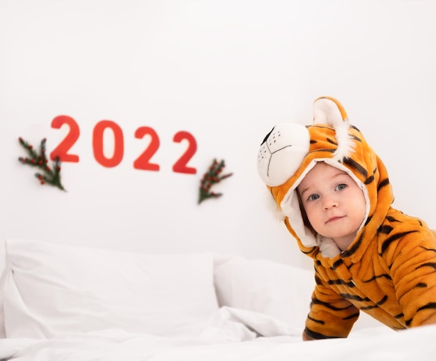 Bebê, criança, criança, criança, criança, caucasiano, sorrindo, vestindo fantasia de tigre na cama branca em casa, olhe para a câmera no fundo dos números 2022. Símbolo do calendário chinês oriental de ano novo.