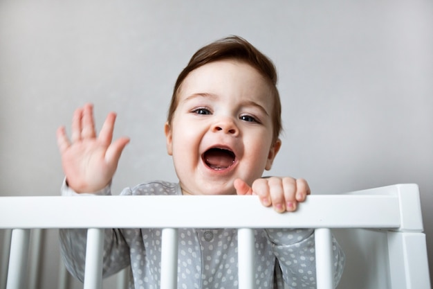 Bebê curioso gritando e de pé em uma cama branca do berço.