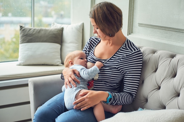 Bebê de 8 meses comendo leite da mãe. Mãe amamentando menino infantil, localização no interior do sofá em casa