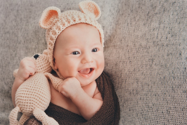 Bebê de olhos claros sorrindo. Uma criança em um boné bege com orelhas e um brinquedo bege de malha na mão