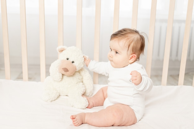 Bebê de seis meses brincando em um berço em um macacão branco com um ursinho de pelúcia