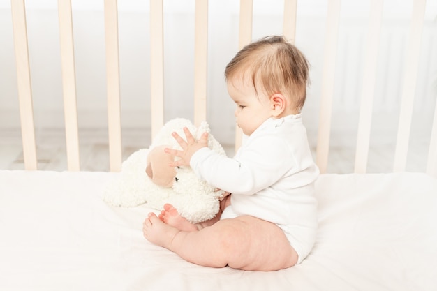 Bebê de seis meses brincando em um berço em um macacão branco com um ursinho de pelúcia