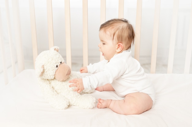 Bebê de seis meses brincando em um berço em um macacão branco com um ursinho de pelúcia