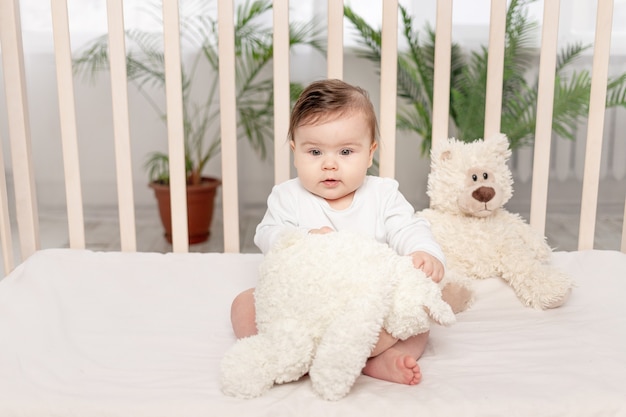 Bebê de seis meses brincando em um berço em um macacão branco com um ursinho de pelúcia