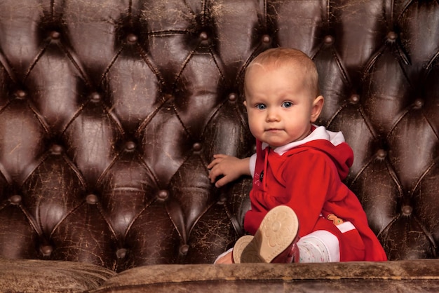Bebê disfarçado de Papai Noel sentado no velho sofá no fundo decorado da árvore de Natal olhando para a câmera