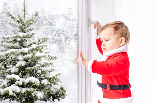 Un bebé con un disfraz de Papá Noel mira y abre la ventana en invierno en un árbol de Navidad cubierto de nieve, el concepto de año nuevo y Navidad