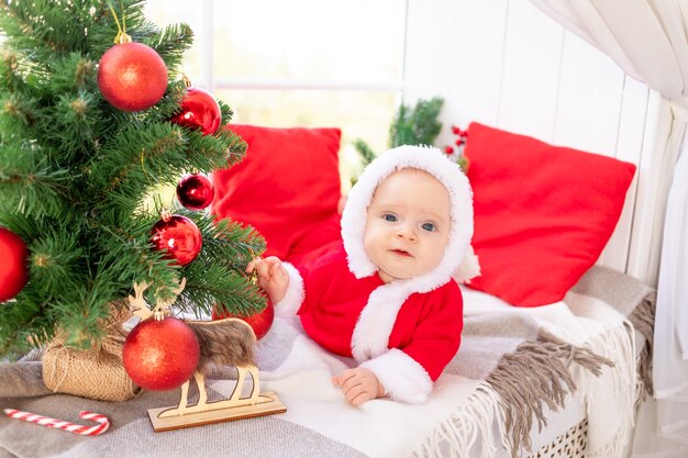 Un bebé disfrazado de Papá Noel bajo el árbol de Navidad juega con bolas rojas de Navidad en la ventana de la casa, el concepto del año nuevo