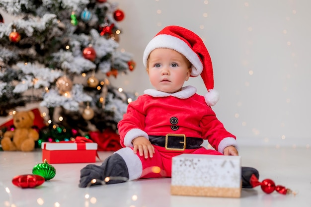 Bebé disfrazado de Papá Noel está sentado al lado del árbol de Navidad y jugando con cajas de regalo. niño abre un regalo de Navidad. concepto de año nuevo de invierno. espacio para texto. foto de alta calidad