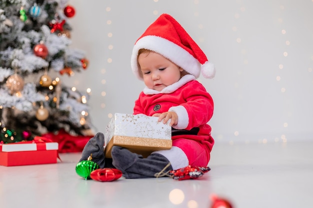 Bebé disfrazado de Papá Noel está sentado al lado del árbol de Navidad y jugando con cajas de regalo. niño abre un regalo de Navidad. concepto de año nuevo de invierno. espacio para texto. foto de alta calidad