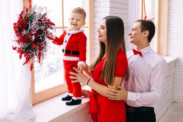 Bebé disfrazado de santa en el alféizar de la ventana padres en ropa festiva junto a la ventana y el bebé corona de Navidad.