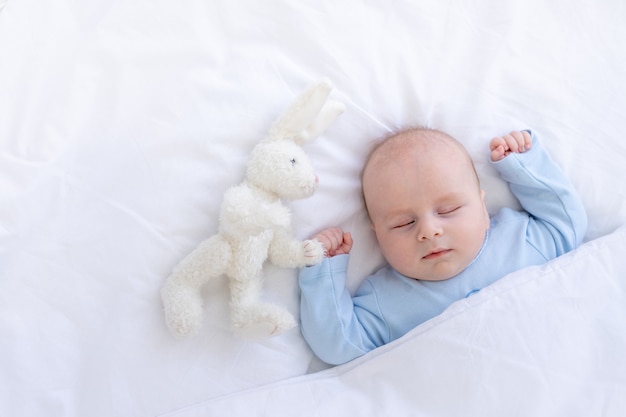 El bebé duerme en la cama acostado de espaldas con un oso de peluche en pijama azul sosteniendo sus manos en alto, un sueño recién nacido saludable