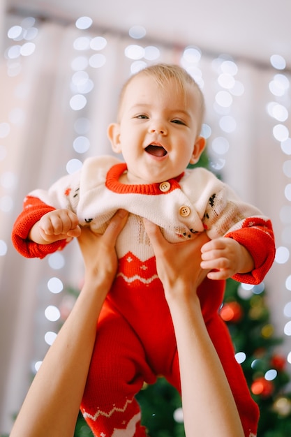 Bebê em um macacão de tricô de Natal nos braços da mãe na frente de uma árvore de Natal. Foto de alta qualidade