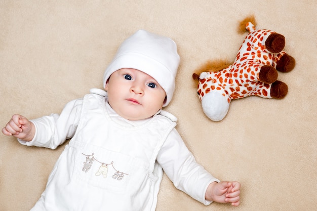 Bebê em um terno branco deitado de costas sobre um tapete bege, ao lado de um brinquedo macio