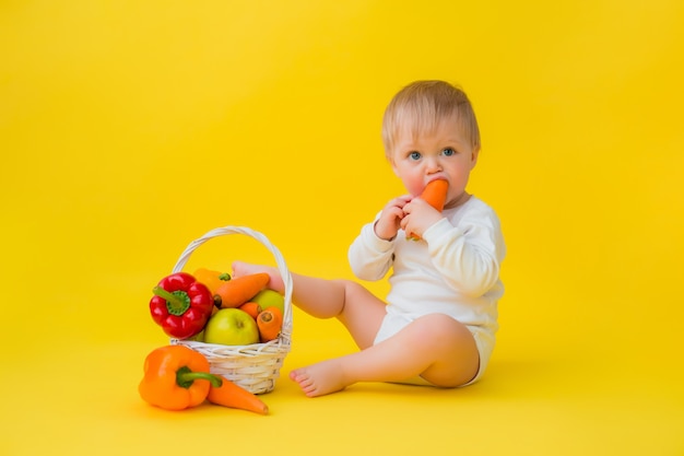 Bebê em uma roupa branca com legumes, sentado sobre um fundo amarelo. comida saudável para a criança, espaço para texto