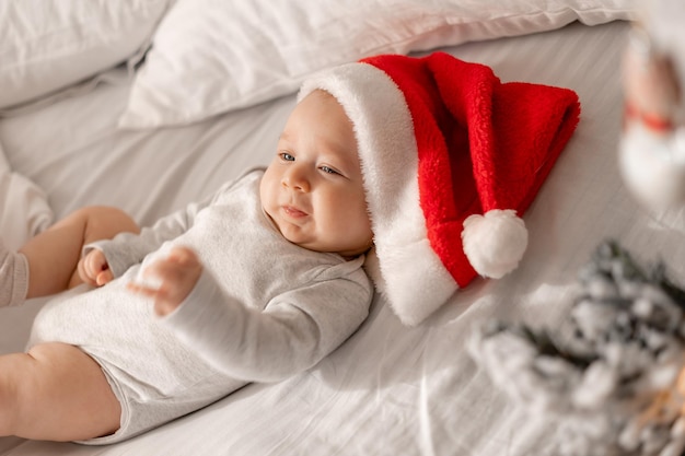 Bebê em uma roupa branca e um chapéu de Papai Noel está deitado na cama ao lado da árvore de Natal. encantadora criança de olhos azuis. férias de inverno, momentos engraçados, natal infantil. espaço para texto. Foto de alta qualidade