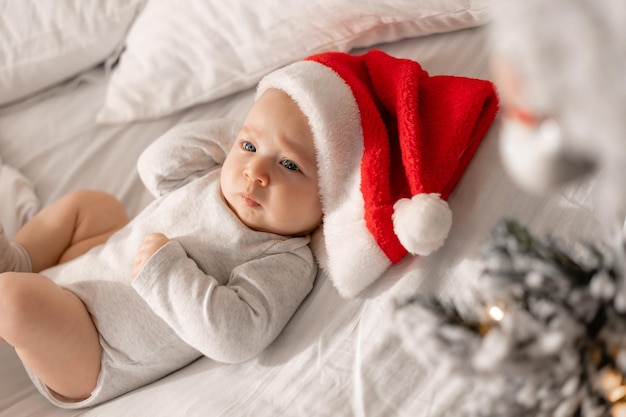 Bebê em uma roupa branca e um chapéu de Papai Noel está deitado na cama ao lado da árvore de Natal. encantadora criança de olhos azuis. férias de inverno, momentos engraçados, natal infantil. espaço para texto. Foto de alta qualidade