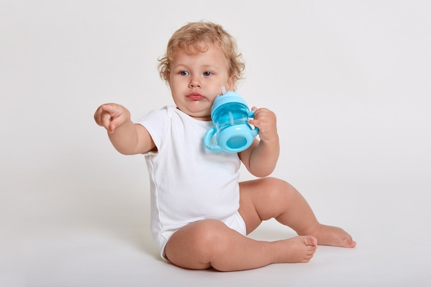 Bebê encantador quer beber água da mamadeira, olhando para longe, apontando com o dedo indicador