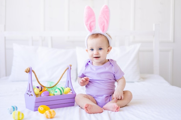 Bebê engraçado bonito com orelhas de coelho com ovos de páscoa coloridos e uma cesta de ovos em casa em uma cama de cama branca cartão de páscoa