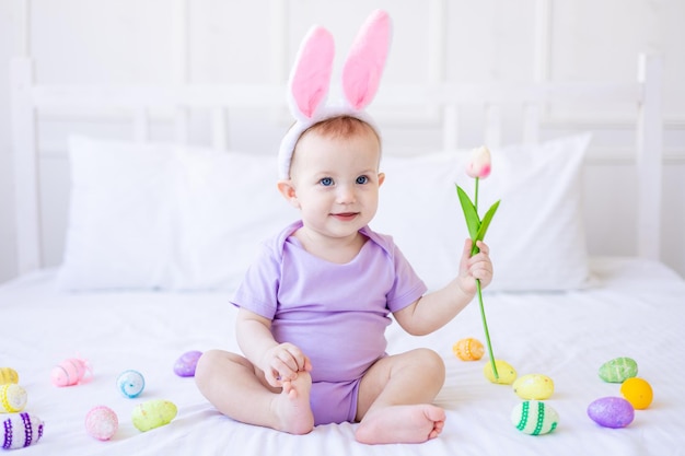 Bebê engraçado bonito com orelhas de coelho com uma tulipa na mão e ovos de páscoa coloridos em casa em uma cama de cama branca cartão de páscoa