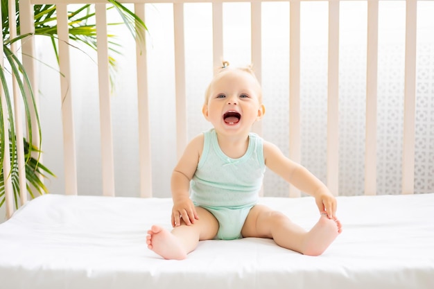 Bebê engraçado de pé em um berço em um berçário branco com uma janela uma garotinha em uma roupa de algodão bebê sorridente de olhos azuis