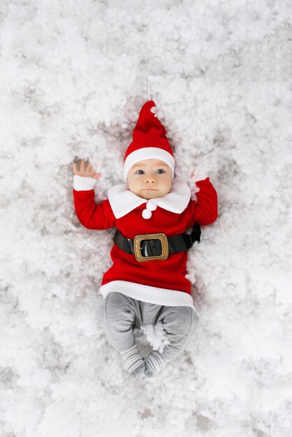 Bebê engraçado engraçado na fantasia de Papai Noel encontra-se na neve artificial.