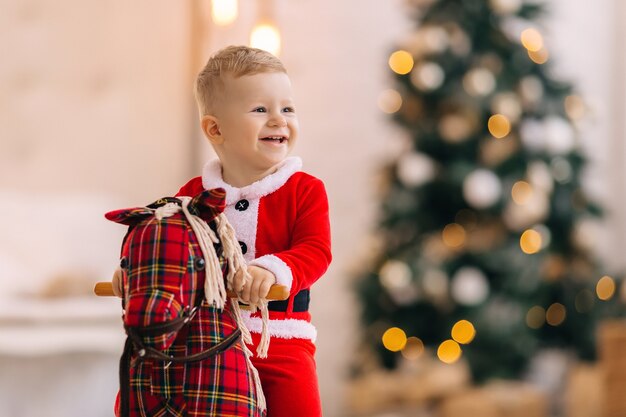 Bebê fantasiado de Papai Noel no cavalo de balanço. árvore de natal borrada