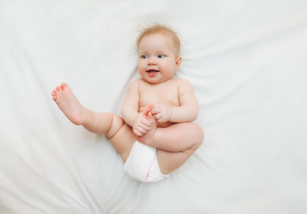 Un bebé feliz y alegre en pañales está acostado en una cama blanca y sosteniendo su pierna Foto de alta calidad para texto