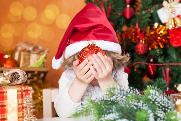 Bebê feliz com chapéu de Papai Noel se escondendo atrás de uma bola contra a árvore de Natal com enfeites