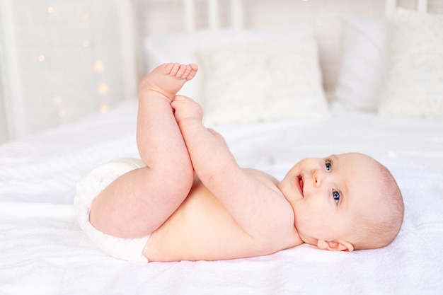 Bebê feliz e rindo brincando de pés em fraldas em um berço em uma cama de algodão branco higiene e cuidados com o recém-nascido