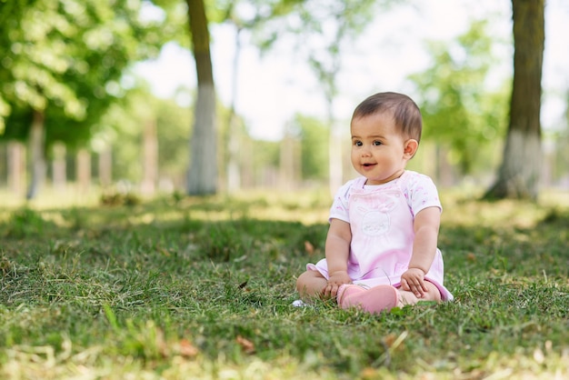 Bebê feliz em um vestido rosa senta-se na grama do parque. Mulher pequena do bebê que plaing na grama verde.