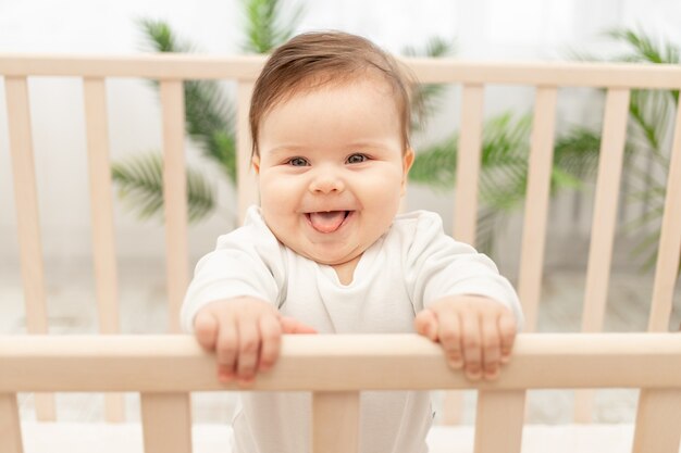 Bebé feliz muestra la lengua mientras está de pie en la cuna