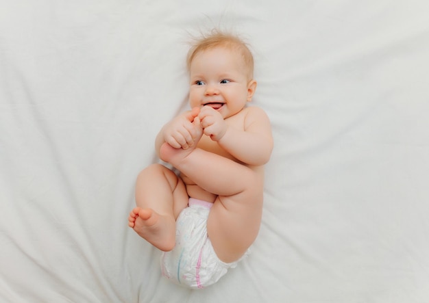 Un bebé feliz en pañales está acostado en una cama blanca y sosteniendo su pierna Foto de alta calidad para texto