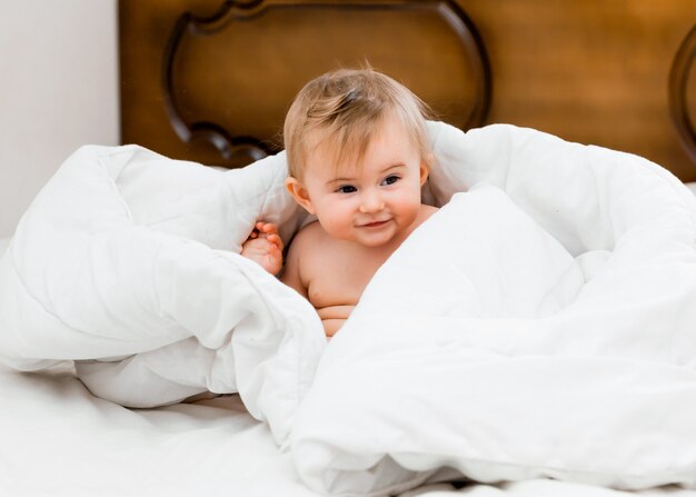 Bebé feliz y saludable de 11 meses sentado en la cama bajo una manta blanca y riendo. concepto de bebe