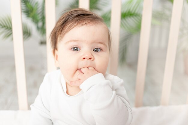 Bebé feliz sentado en la cuna con un traje blanco y chupándose el dedo