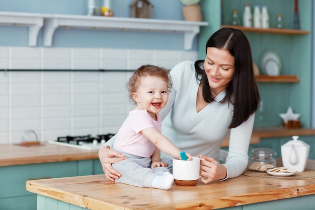 Bebé feliz sentado en la mesa de la cocina con mamá revolviendo gachas con una cuchara