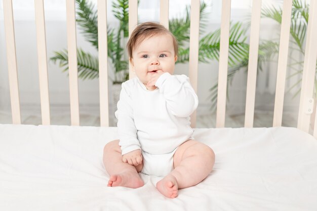 Bebê feliz sentado no berço com uma roupa branca e chupando o dedo