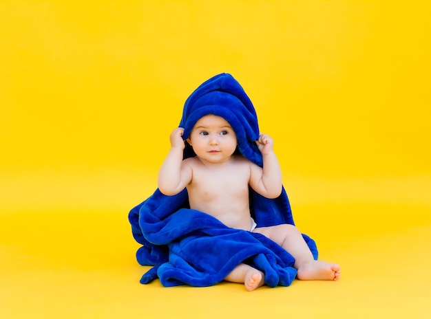 Bebé feliz sentado sobre una superficie amarilla, envuelto en una toalla azul con capucha. bebé después del baño.