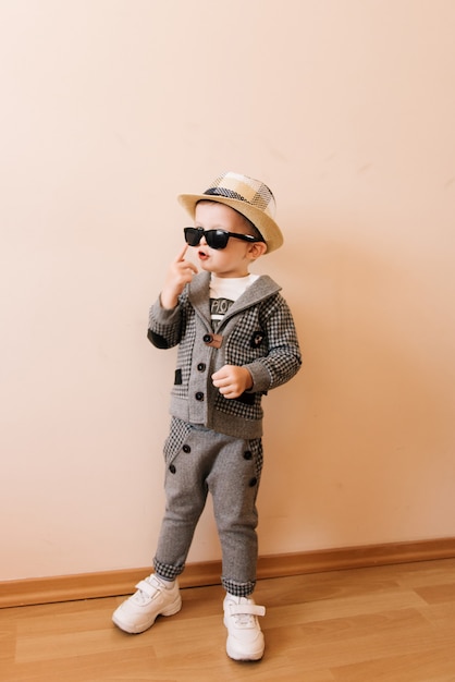 Bebé feliz en traje gris, sombrero y gafas en la pared de luz