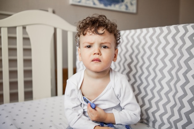 Bebé con fiebre. El niño es un niño pequeño con un termómetro debajo del brazo sentado en la cama. Enfermo, triste