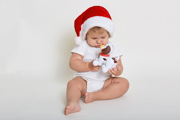 Bebê fofo com chapéu de natal e macacão branco brincando com cachorro de brinquedo de plástico enquanto está sentado descalço no chão