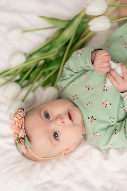 Bebê fofo com uma faixa floral e um body de algodão verde em casa em uma cama branca com tulipas. primavera