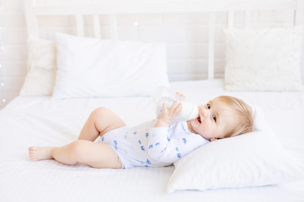 Bebê fofo com uma garrafa de leite é uma merda segurando-o nas mãos, deitado de costas em uma cama de bebê em um conceito de comida para bebê de quarto brilhante para crianças