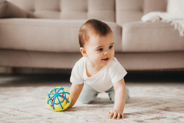 Bebê fofo de 9 meses brincando com uma bola colorida em um berçário com móveis brancos