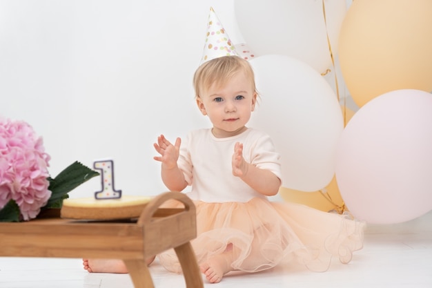 Bebê fofo e feliz, caucasiano, olhando para a câmera, sentado em casa, perto de balões festivos, bolo de flores