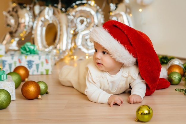 Bebê fofo e sorridente está deitado sob uma árvore de Natal festiva e brincando com presentes. Celebrações de Natal e Ano Novo.